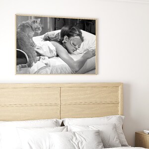 May include: A black and white photograph of a woman sleeping in bed with a cat sitting on the bed beside her. The woman is wearing a sleep mask and the cat is looking at the camera.