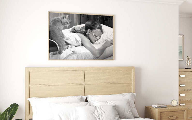 May include: A black and white photograph of a woman sleeping in bed with a cat sitting on the bed beside her. The woman is wearing a sleep mask and the cat is looking at the camera.