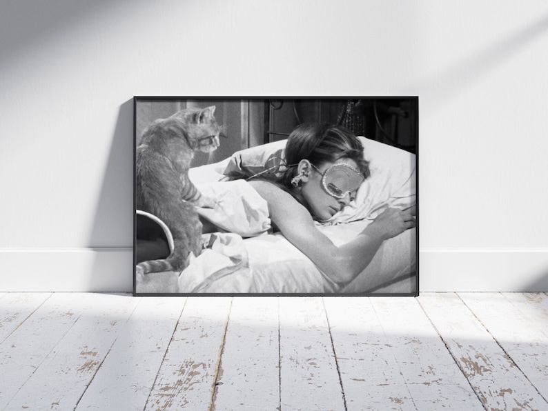 May include: A black and white photograph of a woman sleeping in bed with a cat sitting on the edge of the bed. The woman is wearing a sleep mask and has her hair pulled back. The cat is looking at the camera.
