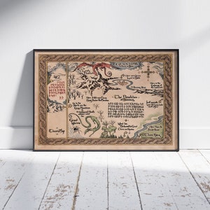 May include: A framed fantasy map with a black border, featuring illustrations of a dragon, mountains, and a river. The map is in shades of brown and beige, with text in a stylized font. The map is leaning against a white wall.