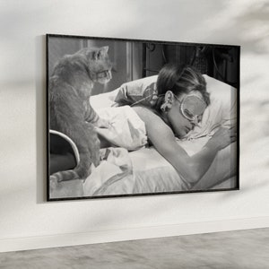 May include: Black and white photo of a woman sleeping in bed with a cat. The woman is wearing a sleep mask and has her hair pulled back. The cat is sitting on the bed next to her.