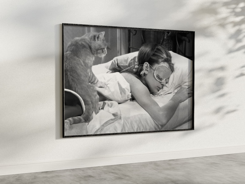 May include: Black and white photo of a woman sleeping in bed with a cat. The woman is wearing a sleep mask and has her hair pulled back. The cat is sitting on the bed next to her.