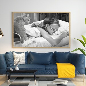 May include: A black and white photograph of a woman sleeping in bed with a cat sitting on the bed behind her. The woman is wearing a sleep mask and has her arms wrapped around a pillow. The cat is looking at the camera.