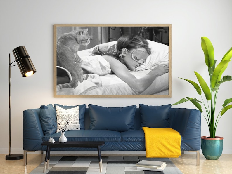 May include: A black and white photograph of a woman sleeping in bed with a cat sitting on the bed behind her. The woman is wearing a sleep mask and has her arms wrapped around a pillow. The cat is looking at the camera.