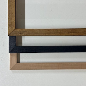 May include: Three wooden picture frames in different colors: dark brown, black, and light brown. The frames are stacked on top of each other, showcasing their various finishes and styles. The frames are displayed against a white background.