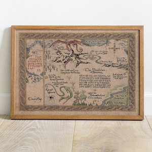 May include: A framed map print with a detailed, fantasy-themed design. The map features mountains, rivers, and text in a stylized font, with a dragon illustration. The frame is made of light brown wood.