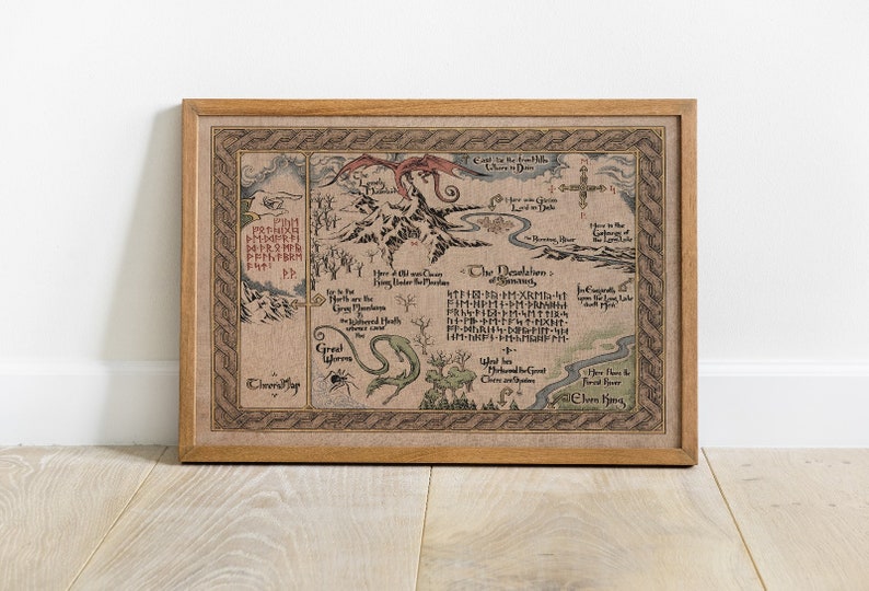 May include: A framed map print with a detailed, fantasy-themed design. The map features mountains, rivers, and text in a stylized font, with a dragon illustration. The frame is made of light brown wood.