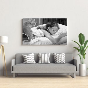 May include: Black and white photo of a woman sleeping in bed with a cat sitting on the bed. The woman is wearing a sleep mask and the cat is looking at the camera.