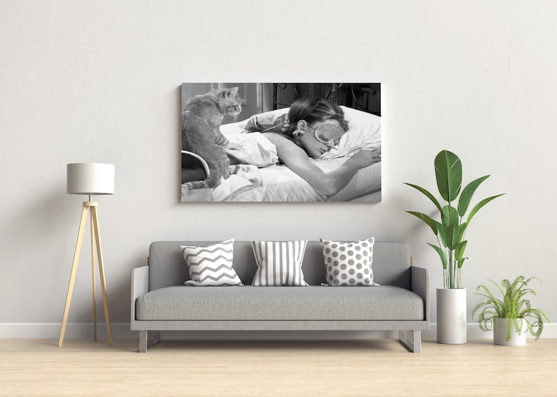 May include: Black and white photo of a woman sleeping in bed with a cat sitting on the bed. The woman is wearing a sleep mask and the cat is looking at the camera.