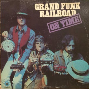 May include: Album cover for Grand Funk Railroad's "On Time." The cover features three men holding toy trains and a pocket watch. The band's name is in white block letters above a purple rectangle with "ON TIME" in white. The Capitol Records logo is in the bottom left corner.