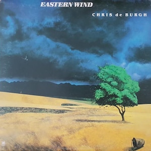 May include: Album cover for "Eastern Wind" by Chris de Burgh. The artwork features a landscape with a green tree, a yellow field, and a dark, cloudy sky. The album title and artist's name are at the top of the cover.