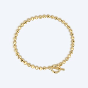 May include: A gold-colored beaded necklace with a toggle clasp. The necklace is composed of small, evenly spaced spherical beads. The clasp features a circular ring and a bar closure. The jewelry is displayed against a white background.