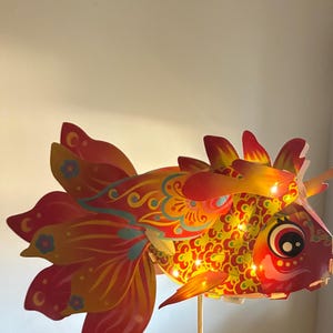 May include: A colorful, decorative fish-shaped lantern with intricate patterns in red, yellow, and orange. The lantern has a series of small lights inside, and is mounted on a wooden stick. The fish has large, detailed fins and tail.