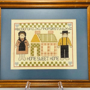 May include: A framed cross-stitch with a blue background. The cross-stitch depicts two figures, a woman and a man, standing in front of two houses. The text "Home Sweet Home" is stitched below the figures. The alphabet is stitched above the figures.