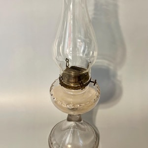 May include: A clear glass oil lamp with a decorative, textured base and a clear glass chimney. The lamp has a brass burner and a metal wick adjuster.