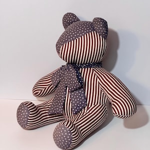 May include: A handmade teddy bear with a patriotic theme. The bear features a striped body in red and white, with gray star-patterned accents on the head, feet, and scarf. The scarf is tied around the neck.
