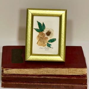 May include: A framed floral art piece with a gold-colored frame, featuring dried flowers and green leaves. The artwork is displayed on top of a stack of vintage, burgundy-colored books. The text "Dear Ladies '95" is visible.
