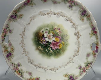 May include: A white porcelain serving plate with a floral design. The plate has two handles and a scalloped edge. The center of the plate features a bouquet of pink, white, and yellow flowers with green leaves. The border of the plate is decorated with a delicate floral pattern in shades of pink, purple, and green.