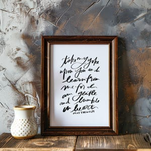 May include: A framed print with the handwritten text "Take my yoke upon you and learn from me for I am gentle and humble in heart. Matthew 11:29." The frame is dark brown. A small white vase with a gold rim sits to the left.