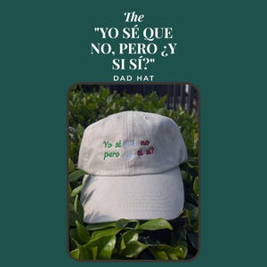 Op de afbeelding: Een beige dad hat met geborduurde tekst op de voorkant die Yo sé que no pero ¿y si sí? leest in groene, blauwe en rode draad. De pet heeft een gebogen klep en een gestructureerde kroon met ventilatiegaatjes op de bovenste panelen.