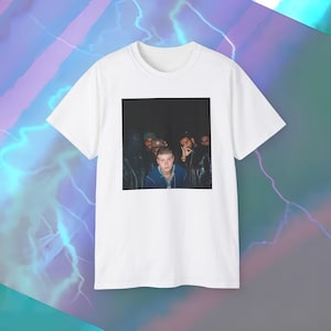 May include: White t-shirt featuring a square image of a group of people. The image shows a person in a blue jacket and a group of people in the background. The t-shirt is made of a soft, comfortable material.