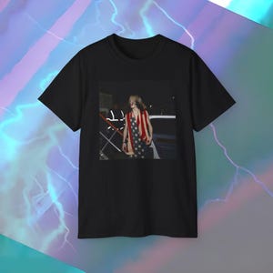 May include: Black t-shirt featuring a photograph of a person wearing a red, white, and blue American flag-themed garment. The image on the shirt appears to be taken at night, with a person and a vehicle visible in the background.