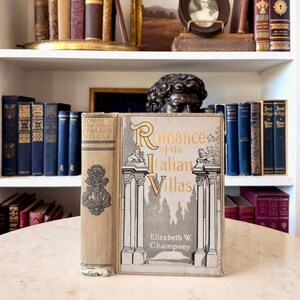 May include: A hardcover book titled Romance of the Italian Villas by Elizabeth W. Champney. The cover features a gray illustration of classical columns and lions with gold lettering. The spine is tan with a decorative black emblem and text.