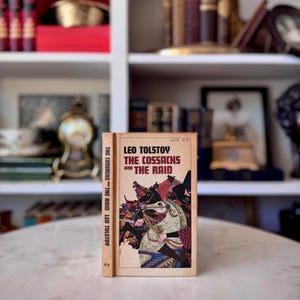 May include: A paperback book titled The Cossacks and The Raid by Leo Tolstoy. The cover features a tan background with a detailed illustration of figures on horseback in shades of red, black, and white. The spine displays the title and author.