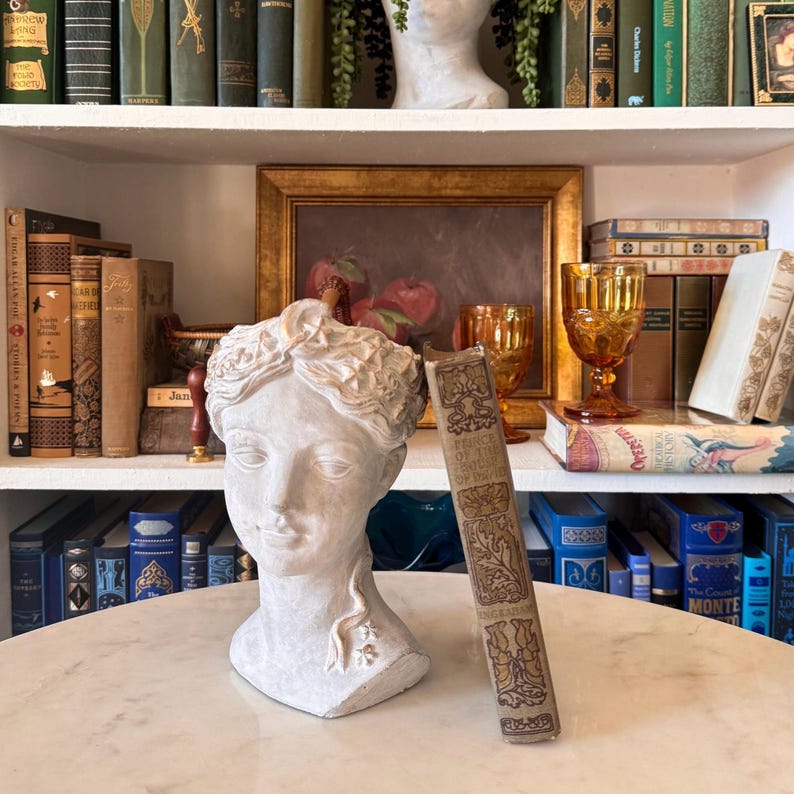 May include: A white plaster bust of a woman and a book titled "Prince of the Gentle People" on a marble table. The book has gold lettering and floral designs. The background includes a shelf of books, a painting, and amber glassware.