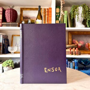 May include: A purple book with the word "ENSOR" in gold lettering. The book is on a marble surface, with a shelf of books and decorative items in the background.