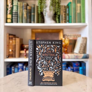 May include: A hardcover book titled "On Writing: A Memoir of the Craft" by Stephen King. The book has a black cover with orange and silver leaf designs and a typewriter illustration. The spine reads "Stephen King On Writing".