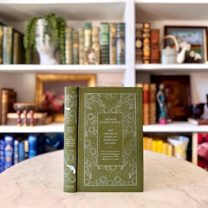 Può includere: Un libro verde con copertina rigida intitolato "The Greatest Cases of Sherlock Holmes" di Arthur Conan Doyle. Il libro presenta dettagli decorativi e testo bianchi. Il dorso è visibile e il libro è in piedi su un tavolo. Un classico.