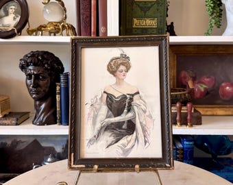 May include: An antique framed print of a woman in a black dress and white gloves, displayed on a decorative gold easel. The print has a dark wooden frame and is set against a backdrop of books and other vintage items.