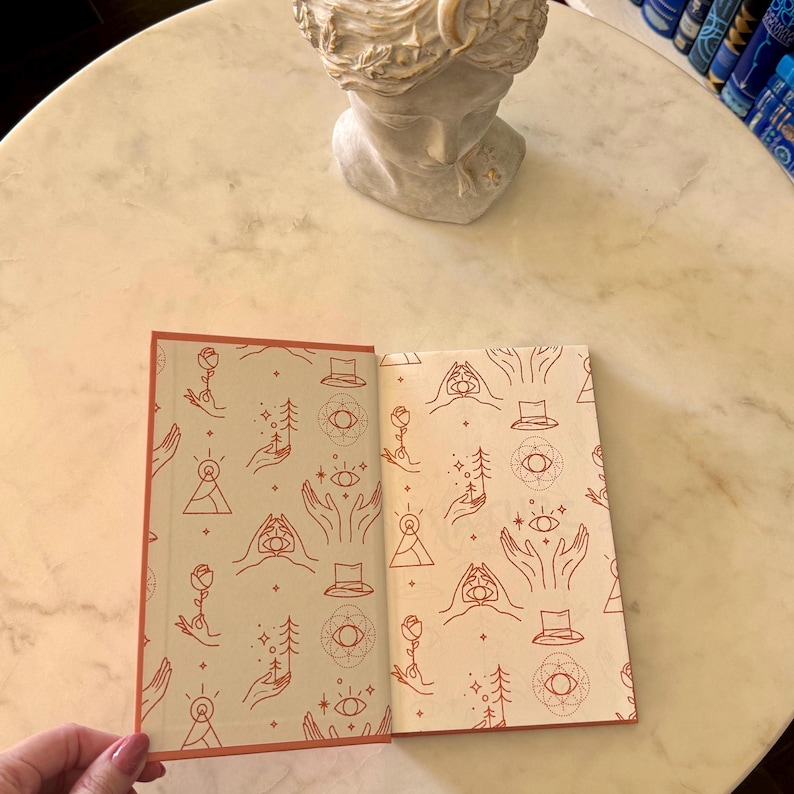 May include: An open book with a cream-colored interior featuring a repeating pattern of hands, eyes, hats, and trees in a reddish-brown color. The book has a reddish-brown border. A marble bust and a stack of books are in the background.