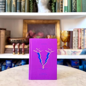 May include: A purple book with gold lettering and a graphic of two blue lightning bolts and a small figure. The book is titled "POETRIES" and "FANCIES OF ATTENTION". The book is on a white table with a blurred background of books and decor.