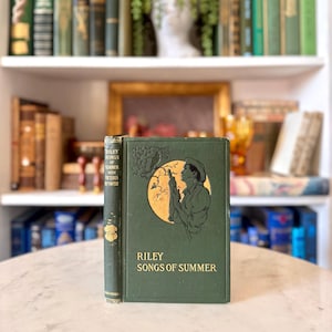 Pode incluir: Dois livros antigos, "Riley Songs of Summer", são exibidos. As capas duras verdes apresentam letras e ilustrações douradas. O livro à direita tem uma imagem circular de um homem. Os livros estão em uma superfície de mármore, com uma estante ao fundo.