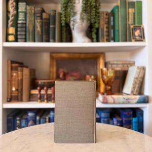 May include: A book with a textured, grey cover stands on a marble table. The background features a white bookshelf filled with antique books in various colours, a framed painting, and decorative items.