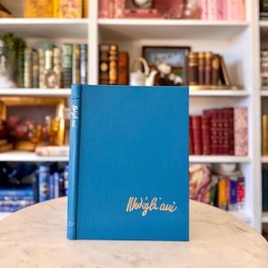 May include: A teal blue book with the title "Modigliani" in gold lettering. The book is standing upright on a marble surface, with a bookshelf in the background. The book's spine also has the title in gold.