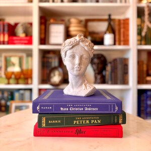 May include: A stack of three classic books with decorative covers, topped by a bust of a woman. The books are titled "Hans Christian Andersen", "Peter Pan", and "Alice". The bust is a light gray color.