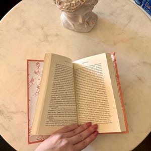 May include: An open book with a red spine and pages, held by a hand. The book's cover features a rose and hand design. The pages are filled with text. The book is on a white marble table, with a bust in the background.