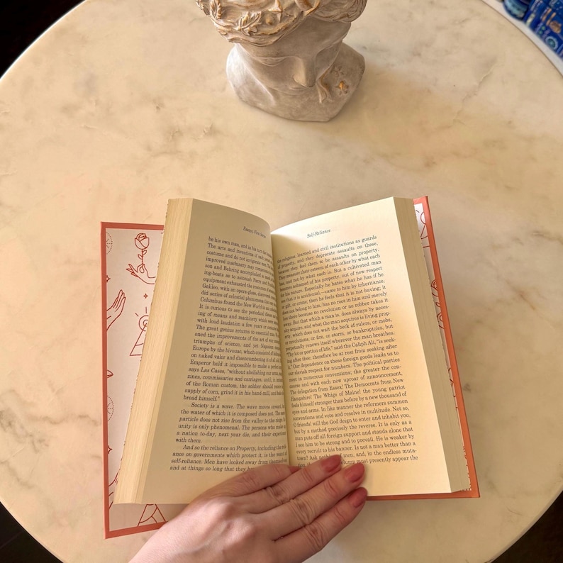 May include: An open book with a red spine and pages, held by a hand. The book's cover features a rose and hand design. The pages are filled with text. The book is on a white marble table, with a bust in the background.