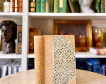 May include: A vintage book with a light tan leather spine and cover. The cover features a patterned design in shades of green, pink, and cream. The book is displayed on a marble surface, with a bookshelf in the background.