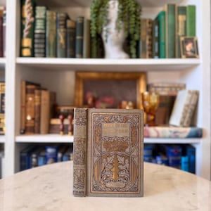 May include: An antique book titled "Prince of the House of David" with gold lettering and decorative accents. The cover is a light grey with gold and brown floral designs. The book is standing upright on a marble surface.