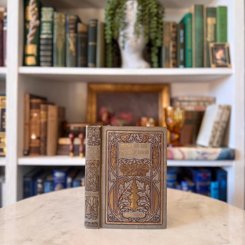 May include: An antique book titled "Prince of the House of David" with gold lettering and decorative accents. The cover is a light grey with gold and brown floral designs. The book is standing upright on a marble surface.