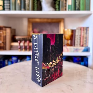 May include: A Little Life book with a black cover depicting a city scene with pink and yellow highlights. The spine shows the title in white script. The book is on a marble table with a blurred background of bookshelves.