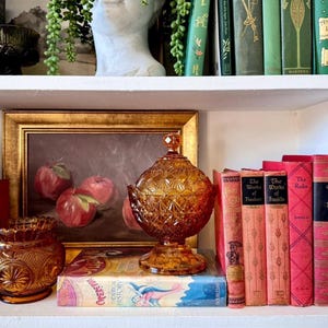 May include: An amber-colored glass candy dish with a diamond-patterned lid and base, positioned on a shelf alongside a framed painting of red apples, a smaller amber glass bowl, and several vintage books with green and red spines.