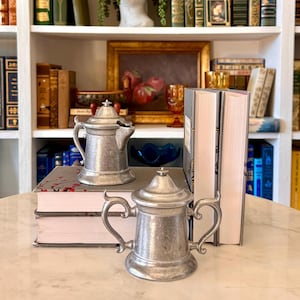 May include: Two antique pewter pitchers with lids, one with a spout, sit on a stack of books. The pitchers have ornate handles and are displayed on a marble surface. A collection of books and vintage decor are in the background.