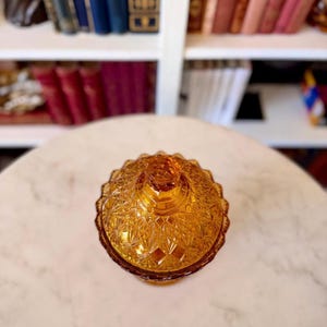 May include: A round amber-colored glass candy dish with a matching lid, featuring a detailed pressed diamond and starburst pattern across the surface. The glass container has a scalloped edge and a small knob handle at the top center of the lid.