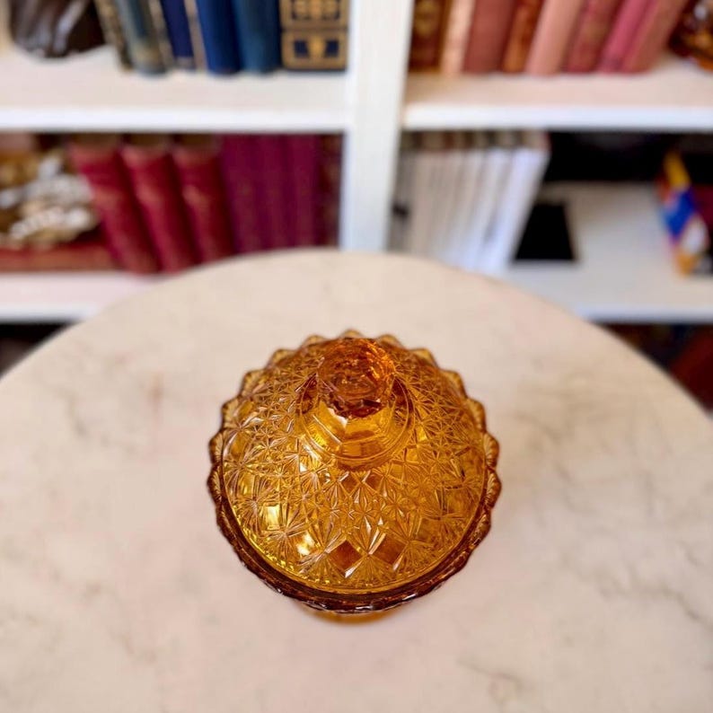 May include: A round amber-colored glass candy dish with a matching lid, featuring a detailed pressed diamond and starburst pattern across the surface. The glass container has a scalloped edge and a small knob handle at the top center of the lid.