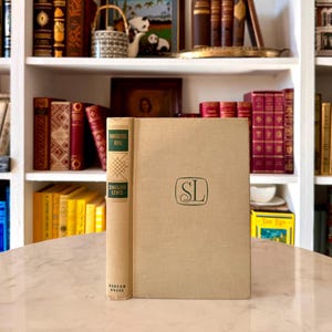 May include: A hardcover book titled "Kingsblood Royal" by Sinclair Lewis, with a beige cover and green lettering. The book is standing upright on a marble surface, with a green "SL" monogram. The background features a bookshelf filled with books.